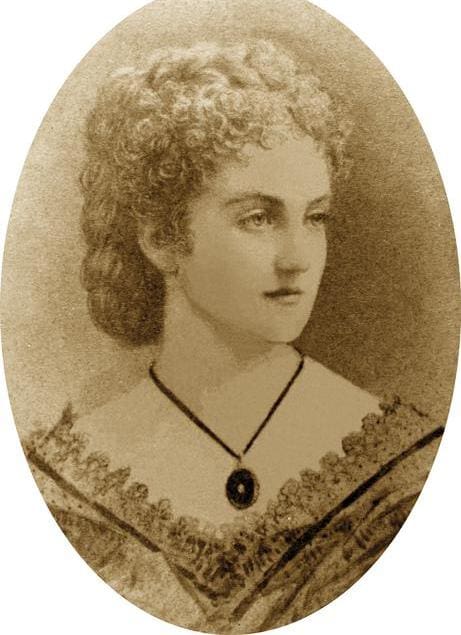 Sepia-toned portrait of a woman with curly hair, wearing an off-the-shoulder dress and a pendant necklace. The image has an oval frame.