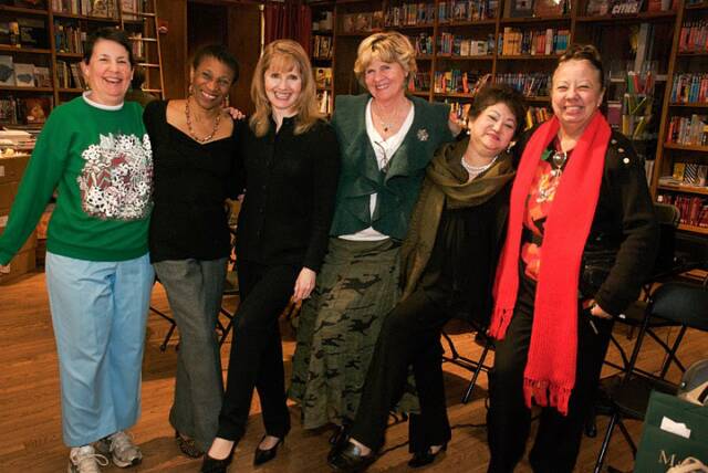 A group of six women stands together, smiling, in a room filled with bookshelves. They are dressed in various casual and semi-formal outfits.