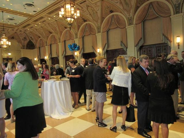 A group of people in formal attire are socializing and mingling in an ornate hall with chandeliers and arched ceilings. Some stand near tall round tables, and there are blue balloons in the background.