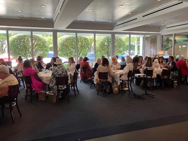 A group of people seated at several tables in a well-lit restaurant with large windows and greenery outside.