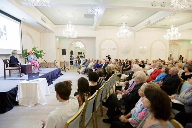 A group of people seated in a chandelier-lit room listens to a speaker on stage during a presentation.