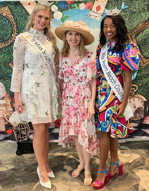 Three women posing together, two wearing sashes that read "Miss South Florida" and "Miss Palm Beach County." The woman in the middle is in a pink floral dress. All are standing in front of a decorative backdrop.