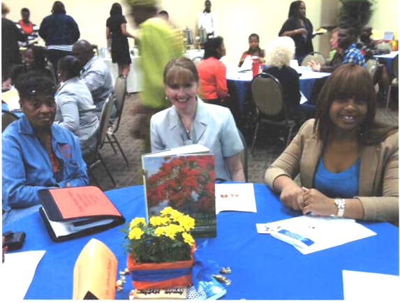 Three women sitting at a blue-covered table with documents and a book centerpiece, surrounded by other attendees at a social event.