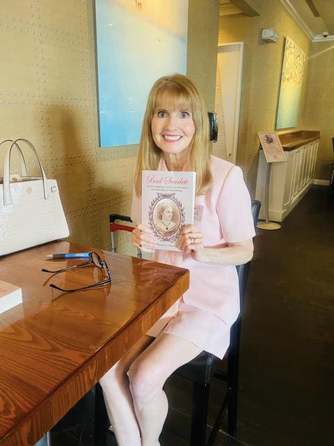 A woman in a pink outfit sits at a table in a café, holding up a book with a smile. On the table are a white purse, glasses, a pen, and another book.