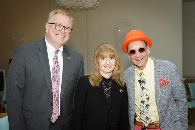 Three people stand together, dressed professionally. The man on the left wears a suit and glasses, the woman in the center has long hair and wears a black outfit, and the man on the right wears a hat and colorful clothes.