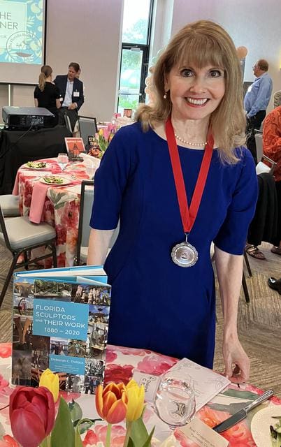 A woman in a blue dress stands at a table with a book titled "Florida Sculptors and Their Work 1860-2020." She is wearing a silver medal around her neck and is smiling at the camera.