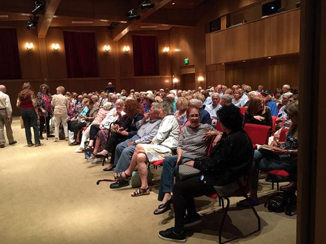 A crowded auditorium with people seated and some standing, engaging in conversations before an event starts.