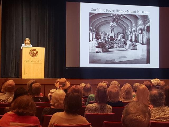 A woman speaks at a podium in front of an audience, with a projected slide showing an image of the Surf Club Foyer at the HistoryMiami Museum.