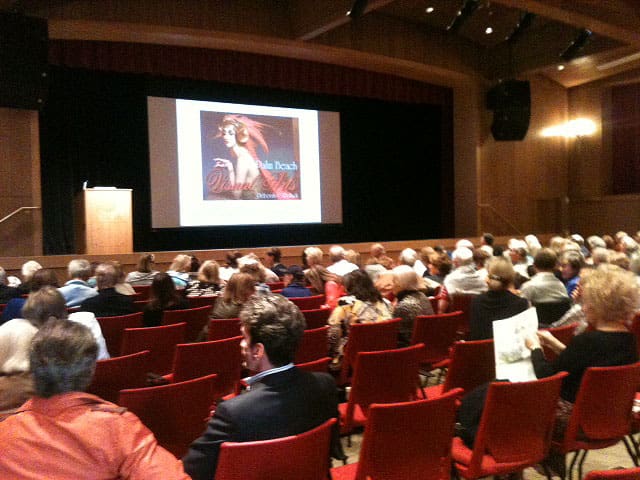 Audience seated in a theater facing a large projector screen displaying an image of a book cover on stage.