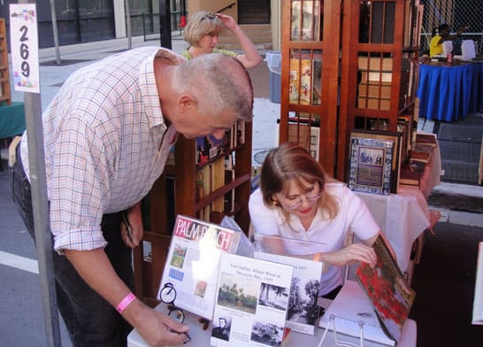 A man and a woman engage in conversation at a book fair booth. The woman is seated, showing the man an open book. Various books and posters are displayed on the table and in the background.