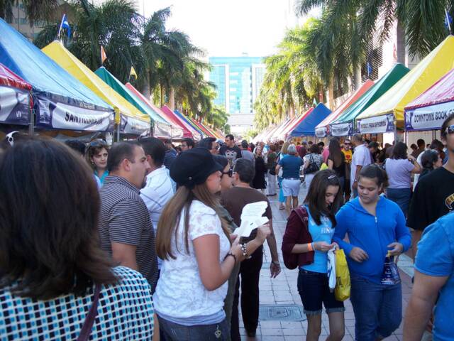 A crowded outdoor market with people walking between colorful vendor tents under palm trees.