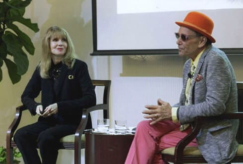 Two individuals sitting on chairs, engaged in conversation. The person on the right is wearing an orange hat, sunglasses, a grey blazer, and pink pants. A projected screen is visible in the background.