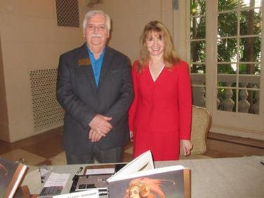 Two people stand behind a table with books and promotional materials. The man is wearing a blue shirt and dark suit jacket; the woman is in a red dress. A window and foliage are visible in the background.