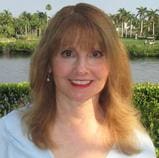 A woman with shoulder-length, auburn hair is smiling at the camera. She is standing outdoors near water with palm trees and greenery in the background.