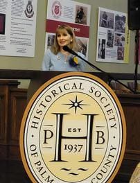 A woman stands at a podium with the logo of the Historical Society of Palm Beach County, delivering a speech. There are informational boards on the wall behind her.