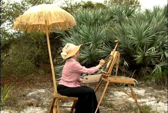 A person wearing a hat and seated on a wooden chair is painting on an easel in an outdoor setting with lush greenery. The setup includes a straw umbrella for shade.