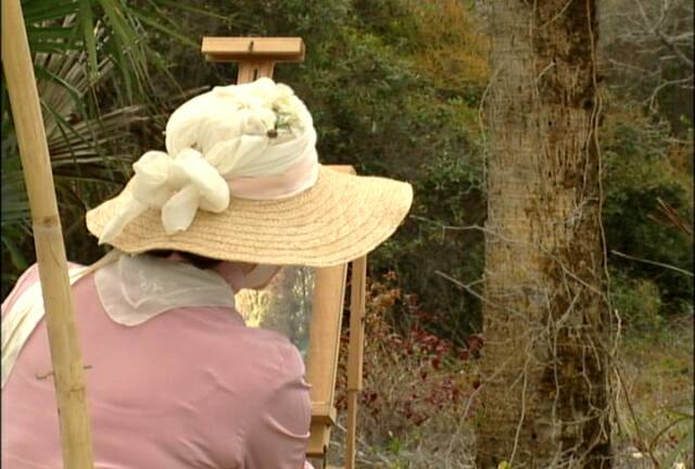 A person wearing a straw hat and a pink outfit paints on an easel outdoors, surrounded by trees.