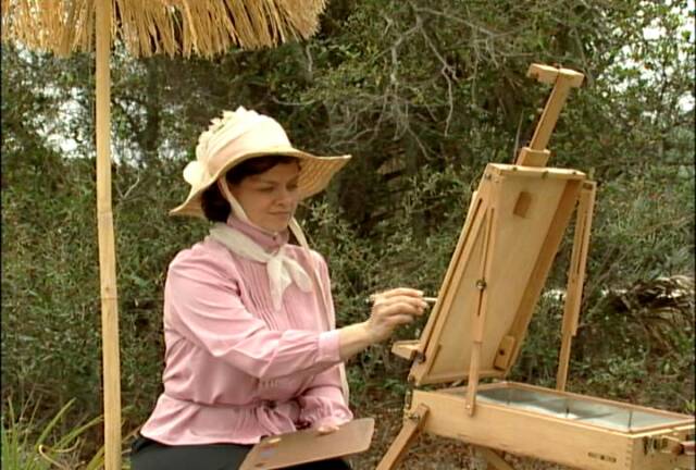 A person wearing a hat and pink outfit is painting on an easel outdoors under a straw umbrella. The person is surrounded by trees and foliage.