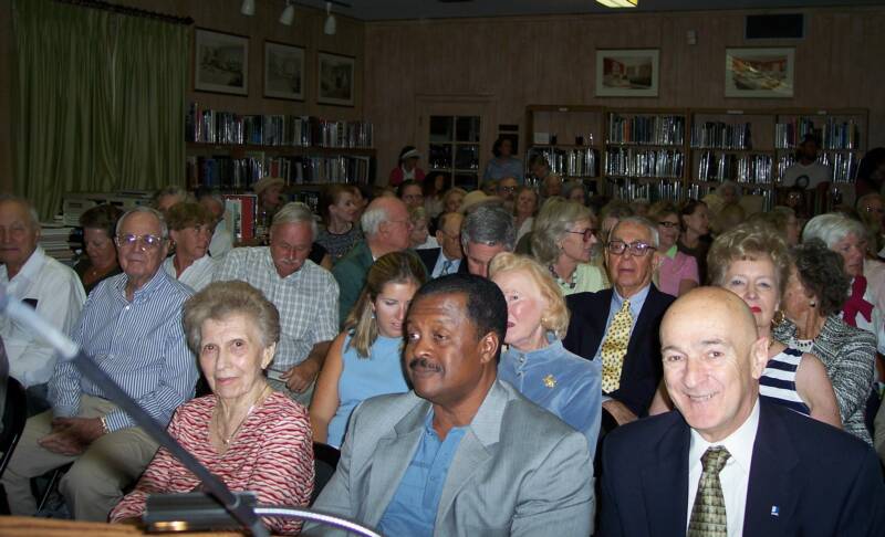 A diverse group of people of various ages seated closely together in a well-lit room with bookshelves in the background.