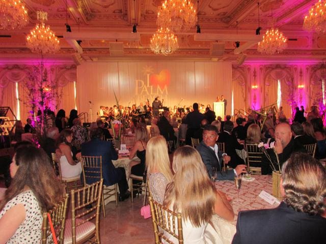 A large crowd attends a formal event in an opulent ballroom with chandeliers and pink lighting. A band performs on stage in the background.