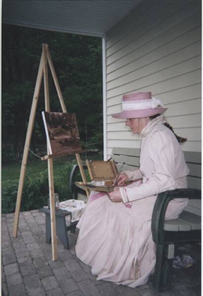 A woman in a pink dress and hat sits on a bench outdoors, painting a landscape on a canvas held by a wooden easel. She is focused on her work, with paint supplies beside her.